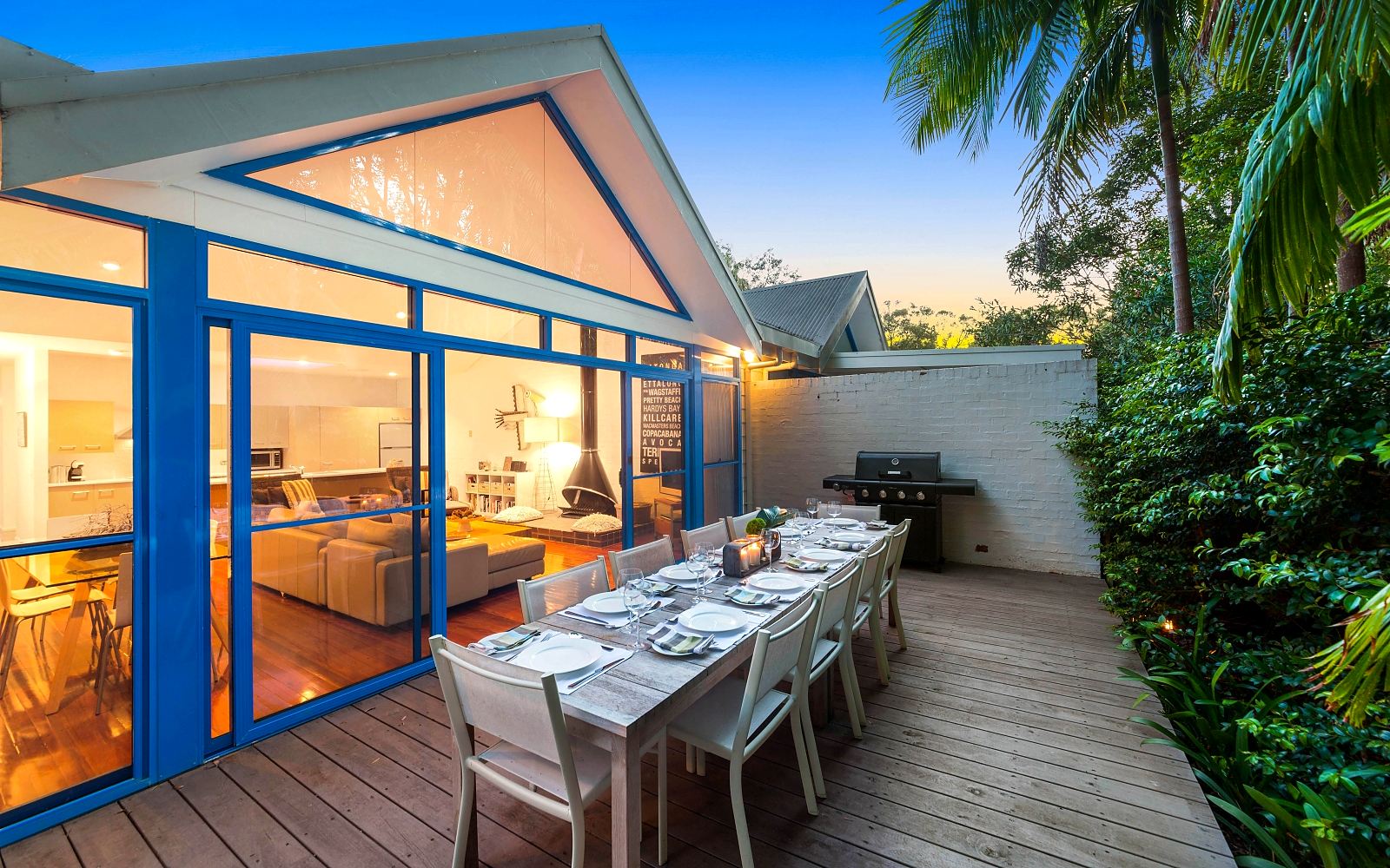 Sails Pearl Beach at dusk: the sail-form roof and blue-framed glass lit from inside, the table set on the deck